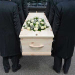 Four people carrying a coffin with white flowers.