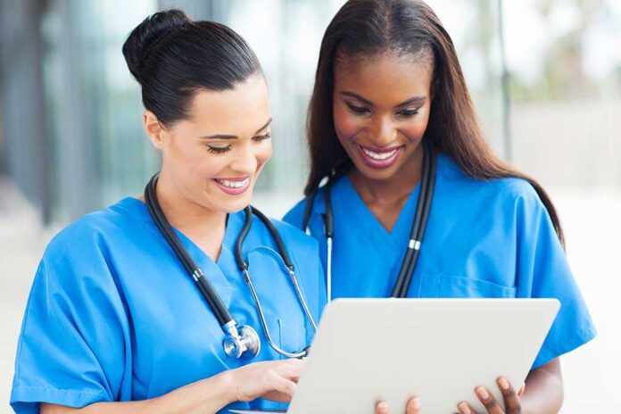 Two nurses in blue scrubs smiling while looking at a tablet
