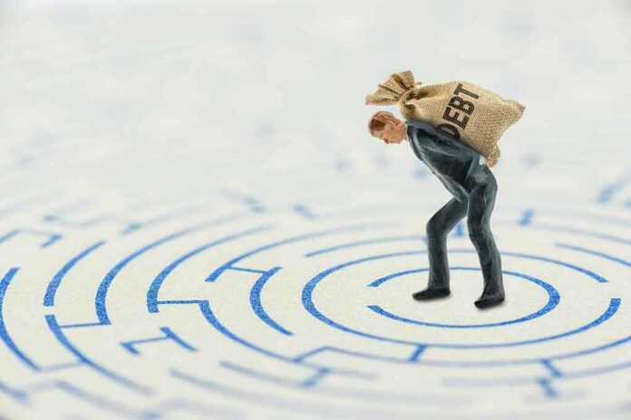 shutterstock_1699344271.jpg A miniature businessman carrying a sack labeled 'DEBT' in a maze