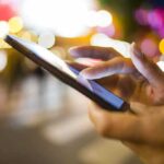 iPhone Horror: 75 Days Wasted Yearly A persons hand using a smartphone in a city at night with blurred lights in the background