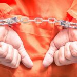 Grave Robber Caught With 100+ Human Skulls Hands in handcuffs wearing an orange jumpsuit