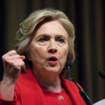 Person speaking passionately at a podium, wearing red jacket.