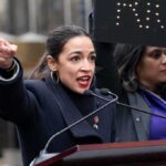 A woman passionately speaking at a rally with a sign in the background