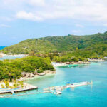 Americans Advised to NOT Travel Here! Tropical beach with docks and lush green hills.