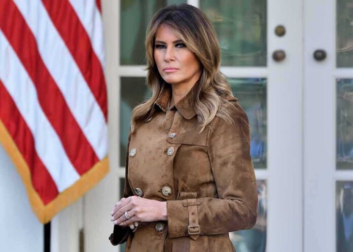 shutterstock_1572261598 Melania Trump standing in a brown suede coat near an American flag