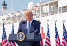 Trump Repeatedly Uses Anti-Trans Slur to Attack Dem Candidate Donald Trump speaking at a podium with American flags and a ship in the background